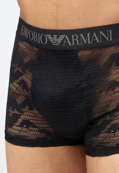 Κοντινό πλάνο ενός ατόμου που φοράει μαύρα δαντελένια μποξέρ της Emporio Armani με ημιδιαφανή γεωμετρικά σχέδια και ελαστική μέση.