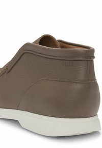 Scarpa in pelle marrone con finitura testurizzata, dotata di suola in gomma bianca. Include un branding sottile sul tallone. Silhouette arrotondata.