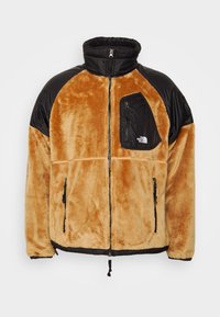 Giacca fuzzy marrone e nera con colletto alto, zip frontale, tasca sul petto con zip e logo, e tasche laterali con zip.