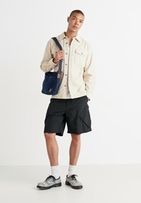 Bēša pogu krekls, melni kravas šorti, tumši zils soma un pelēkas sporta kedas ar baltām zeķēm. Vienkāršs dizains, kokvilnas materiāls, ikdienišķs piegriezums.