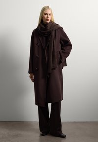 Abrigo de lana marrón oscuro con diseño de doble pecho, grandes botones y una bufanda a juego, llevado sobre pantalones de pierna ancha marrón oscuro.