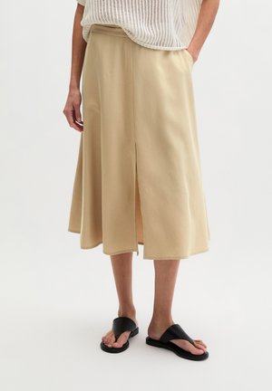 Jupe midi beige avec une fente devant, associée à des sandales noires sans bride et un haut tricoté blanc, jambes et mains partiellement visibles.
