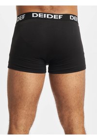 Black boxer shorts made of smooth fabric, with a wide elastic waistband featuring a white logo. Fitted design, mid-thigh length.