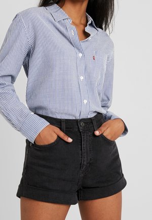Femme portant une chemise à rayures verticales bleues et blanches boutonnée, rentrée dans un short en denim noir taille haute, les mains dans les poches.