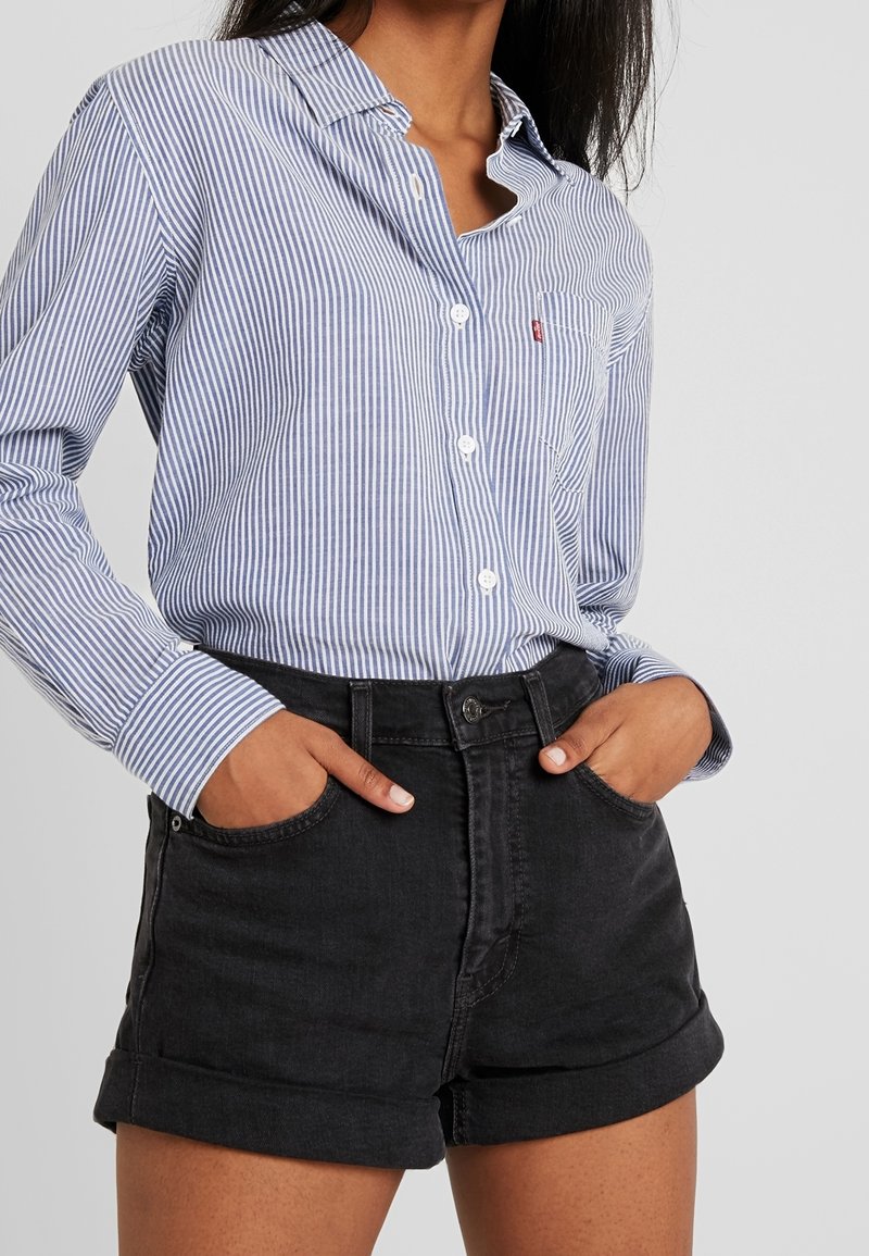 Femme portant une chemise à rayures verticales bleues et blanches boutonnée, rentrée dans un short en denim noir taille haute, les mains dans les poches.