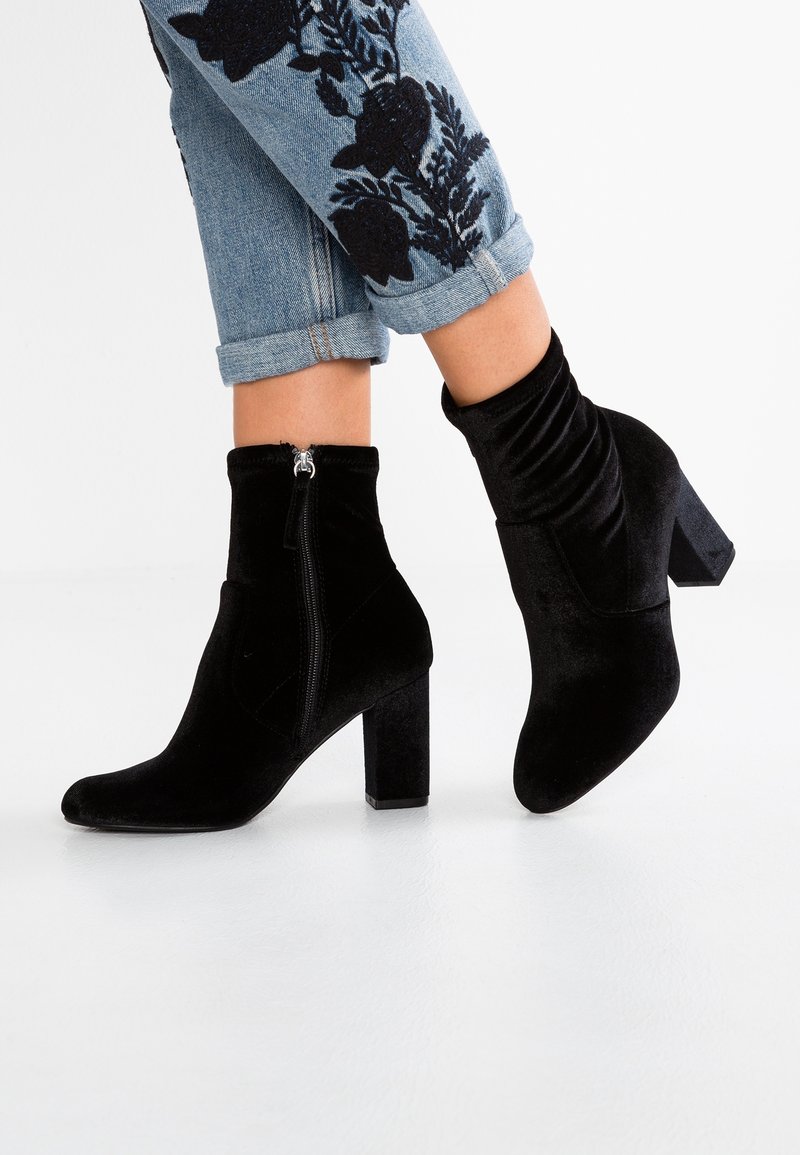 Bottines en velours noir avec talon carré, dotées d'une fermeture éclair latérale et d'une partie supérieure plissée pour la texture, associées à un jean bleu aux revers retroussés.