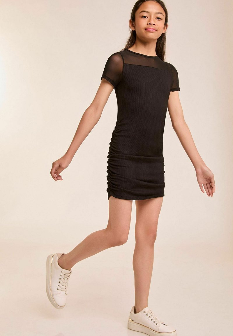 Schwarzes Rippkleid mit kurzen durchsichtigen Ärmeln, ausgestattet mit seitlicher Raffung. Kombiniert mit weißen Sneakers. Der Hintergrund ist hell und neutral.