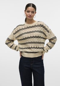 Maglione in pizzo lavorato a maglia color crema con motivi a zigzag neri. Presenta un collo tondo e polsini a costine, abbinati a pantaloni in denim scuro.