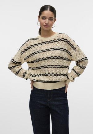 Maglione in pizzo lavorato a maglia color crema con motivi a zigzag neri. Presenta un collo tondo e polsini a costine, abbinati a pantaloni in denim scuro.