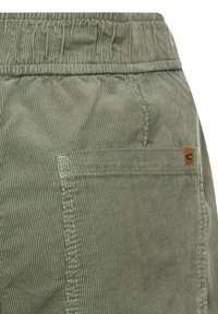 Shorts en velours côtelé gris-vert avec une ceinture élastique, une poche latérale et une petite étiquette logo marron près de la poche.