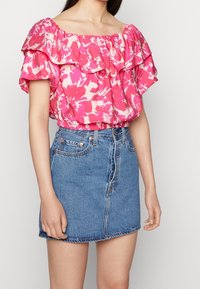 Blouse rose à motifs floraux, épaules dénudées, avec des manches volantées, associée à une jupe en denim ajustée avec fermeture par boutons et ourlet effiloché.