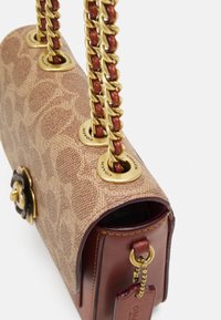 Borsa a tracolla marrone con motivo, dotata di catene dorate infilate con pelle, occhietti dorati incisi con la scritta "COACH" e un'etichetta in pelle attaccata con una catena di perline.