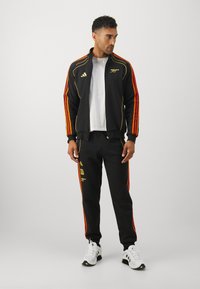 Survêtement noir avec des accents rayés orange et jaune, veste zippée, poignets élastiques et pantalon assorti avec des détails de logo. Baskets blanches.