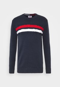 Pull en mailles de couleur marine avec un col rond et des manches longues. Présente une rayure horizontale rouge, blanche et rouge sur la poitrine ainsi que des détails de logo.