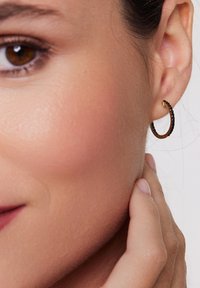 Złota kolczyk w kształcie obręczy z czarnymi kamieniami wzdłuż krawędzi, w zaokrąglonym kształcie i fakturowanej powierzchni, noszony na uchu. W pobliskiej okolicy widoczny odcień skóry.