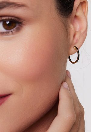 Złota kolczyk w kształcie obręczy z czarnymi kamieniami wzdłuż krawędzi, w zaokrąglonym kształcie i fakturowanej powierzchni, noszony na uchu. W pobliskiej okolicy widoczny odcień skóry.