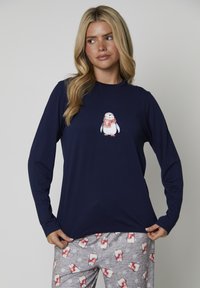 Camicia a maniche lunghe blu navy con grafica di un pinguino con una sciarpa rossa; abbinata a pantaloni da pigiama grigi con motivi di pinguini e fiocchi di neve.