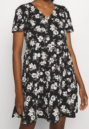 Kobieta w krótkiej, czarnej sukience w kwiaty z białymi i różowymi kwiatami, stojąca na tle jednolitego tła.