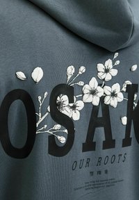 Grå hettegenser med svart tekst "OSAK" og hvite blomsterdesign. Stoffet ser glatt ut med hette. Teksten er trykket under designet.