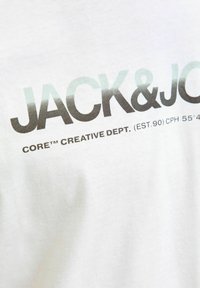 Balta kokvilnas T-kreklā ar drukātu tekstu: "JACK&JONES CORE™ CREATIVE DEPT. (EST.90) CPH 55°43'". Teksts pelēkā un zaļā gradientā.