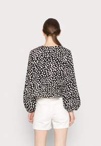 Blusa preta com padrão abstrato branco e lavanda, mangas longas e bufantes, ligeira pregueado na cintura, combinada com calções brancos.