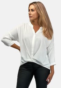 Weiße Bluse mit langen Ärmeln, Knopfleiste und umgeschlagenem Kragen. Glattes Material, lockere Passform und ein V-förmiges Frontdesign. Kombiniert mit schwarzen Hosen.