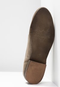 Semelle d'une chaussure plate en daim marron avec un talon en caoutchouc, un logo papillon embossé et le texte "Felmini Falling in Love".