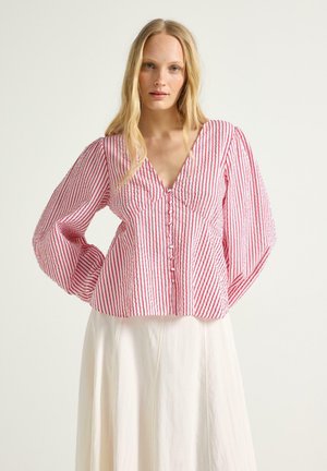 Kvinde iført en rød og hvid bluse med lodrette striber, puffede ærmer og knapper, kombineret med et hvidt høj-taljeret skørt.