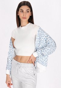 Giacca zip-up leopardata blu chiaro, top fitted bianco e pantaloni sportivi grigi con un dettaglio ricamato "Stay Positive". Texture lisce.