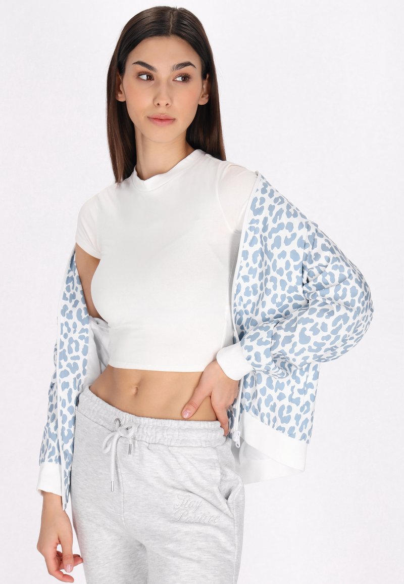 Giacca zip-up leopardata blu chiaro, top fitted bianco e pantaloni sportivi grigi con un dettaglio ricamato "Stay Positive". Texture lisce.