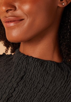 Haut en tissu noir texturé avec un col en volants, complété par de petites boucles d'oreilles en clous et un teint de peau lisse. Accent sur le col et le détail du tissu.