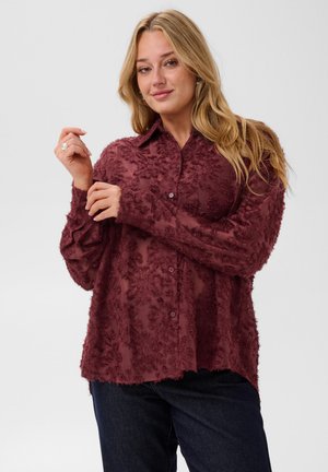 Femme portant une chemise bordeaux texturée à boutons et un jean foncé, ajustant son poignet gauche avec un léger sourire sur un fond blanc.