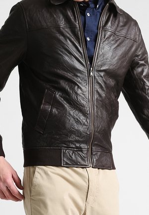 Veste en cuir marron foncé avec fermeture éclair à l'avant, portée sur une chemise bleue et un pantalon beige, sur une personne devant un fond blanc.
