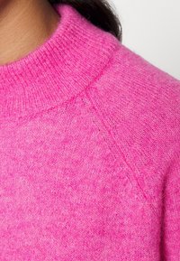 Luddig rosa tröja med ribbad, hög halsringning och raglanärmar. Mjuk textur med en något fläckig utseende, som visar en livlig färg.