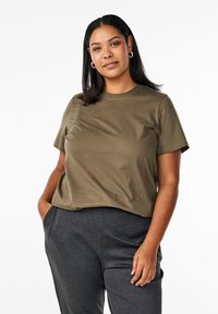 T-shirt in cotone verde oliva con maniche corte, scollatura rotonda, vestibilità rilassata; il modello indossa pantaloni grigio scuro, in piedi con le mani nelle tasche.