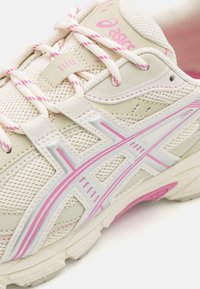 Beige träningsskor med mesh och syntetiskt ovandel, rosa detaljer, reflexstriper och strukturerad sula för grepp. Lättviktsdesign.