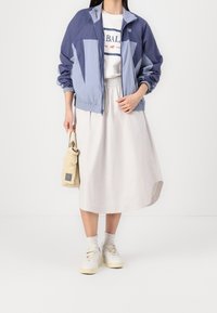 Osoba oblečená v modré zipové bundě, bílé tričko s grafikou, béžové midi sukni a bílých teniskách, držící malou béžovou kabelku.
