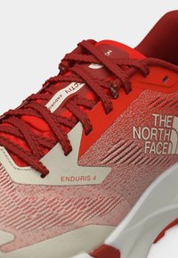 Κόκκινο και μπεζ αθλητικό παπούτσι North Face Enduris 4 με υφασμάτινο επάνω μέρος με υφή και παχιά λευκή σόλα, εμφανίζοντας σφιχτά δεμένα κόκκινα κορδόνια.