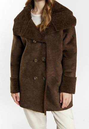 Femme portant un manteau en peau retournée marron à double boutonnage avec un large col et des poignets, par-dessus une chemise blanche et un pantalon clair.
