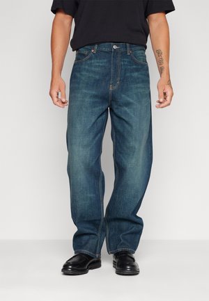 Man wearing loose-fit faded blue jeans, black shoes, and a black shirt, standing with arms relaxed by sides on a plain background.