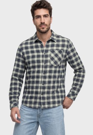 Mann trägt ein schwarz-weiß kariertes Langarmhemd mit Vordertasche und hellblauer Jeans, steht vor einem einfarbigen Hintergrund.