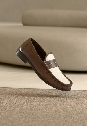 Bruine en witte leren loafer schoen die boven een beige tapijt zweeft met een vervaagde beige bank op de achtergrond.