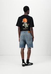 Camiseta negra con estampado floral, pantalones cortos de mezclilla azules con bordes deshilachados y zapatillas negras con acentos blancos, visibles por la espalda.