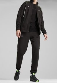 Zwart atletisch trainingspak met een gestructureerd jack met groene accentlijnen, gecombineerd met bijpassende broek en zwarte schoenen.