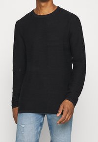 Man wearing a black ribbed long-sleeve shirt and light blue distressed jeans, standing with hands relaxed by sides.