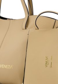 Beżowa skórzana torba o strukturalnym designie, z kontrastującym czarnym przeszyciem oraz małą kieszonką z przodu. Widoczny akcent w postaci złotego logo.