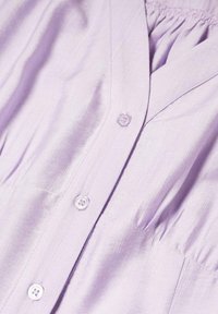 Chemise boutonnée mauve clair avec un tissu texturé, un design plissé et trois boutons visibles près de l'encolure.