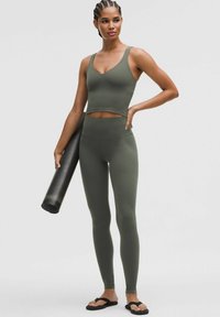 Kvinde i olivengrøn træningscrop-top og leggings, der holder en sort yogamåtte, står med den ene hånd på hoften og iført sorte sandaler.