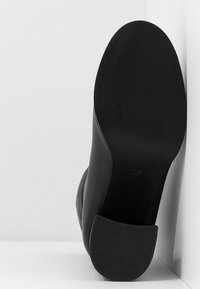 Chaussure noire à talon incliné vers le haut, montrant une semelle texturée et un talon bloc, placée devant un fond blanc en coin.