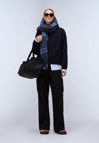 Schwarze Cargo-Hosen, marineblaues Sweatshirt, blauer karierten Schal, dunkle Sonnenbrille und eine schwarze Reisetasche. Lässiges, mehrschichtiges Outfit mit kontrastierenden Texturen.